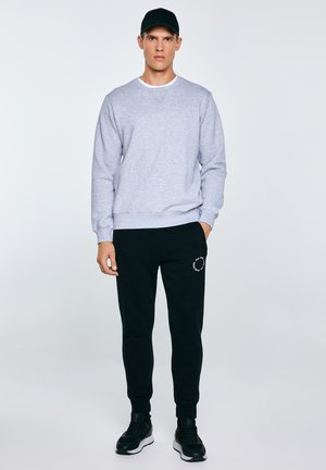 Młody mężczyzna ubrany w szarą bluzę, czarne spodnie dresowe z białym napisem na udzie, czarną czapkę i czarne trampki, stoi z jedną ręką w kieszeni.