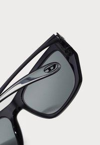 Óculos de sol pretos com armações angulares e lentes cinzas. Apresentam um detalhe metálico nas hastes e um acabamento brilhante.