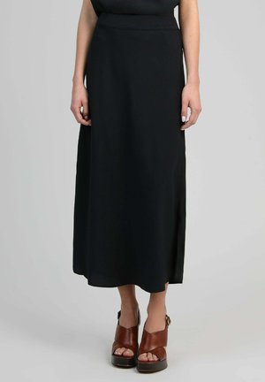 Femme portant une longue jupe noire et des sandales compensées marron à bout ouvert, debout devant un fond uni.