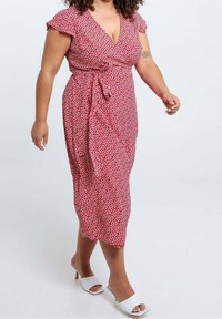 Rotes Blumenkleid mit kurzen, schwingenden Ärmeln, knielang, tailliert und mit kleinen weißen Herzen. Kombiniert mit weißen offenen Absatzschuhen.