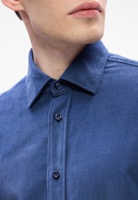 Gros plan d'une personne portant une chemise bleue en velours côtelé à boutons avec un col, montrant la partie inférieure du visage et la zone du cou.