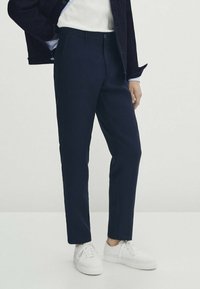 Mörkblå byxor i skräddarsydd passform, tillverkade av ett slätt tyg, med knappstängning fram och snedställda sidofickor. Bärs med vita sneakers.