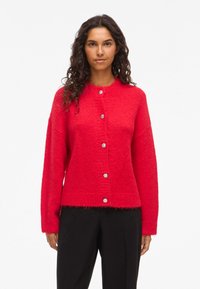 Femme avec de longs cheveux bouclés, portant un cardigan rouge vif à boutons et un pantalon noir, se tenant devant un fond blanc uni.