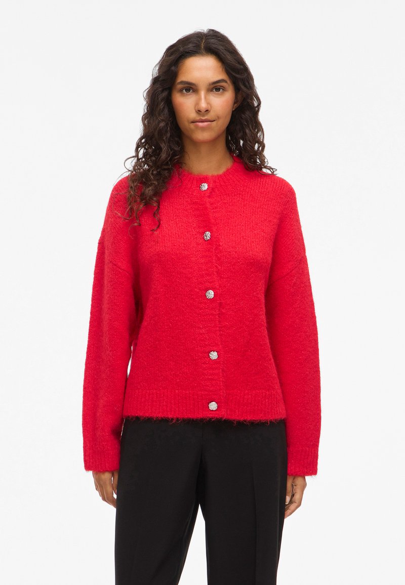 Femme avec de longs cheveux bouclés, portant un cardigan rouge vif à boutons et un pantalon noir, se tenant devant un fond blanc uni.