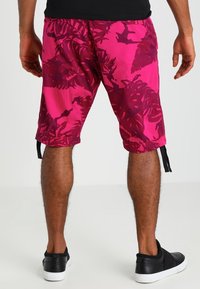 Man som bär ljusrosa tropiska mönstrade shorts, svarta sneakers och en svart t-shirt, visad bakifrån mot en vit bakgrund.