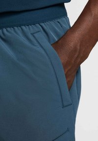 Men's dark teal athletic shorts with an elastic waistband, side pocket, and smooth fabric texture, highlighting a close-up view of the design.
