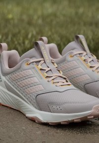 Ανοιχτό γκρι και απαλό ροζ παπούτσια trail running Adidas Terrex με υφασμένες σόλες που ξεκουράζονται πάνω σε τσιμέντο με θολό φόντο χόρτου.
