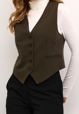 Frau trägt eine dunkelbraune, geknöpfte Weste über einem weißen Langarmrollkragenpullover und schwarzen Hosen, von den Schultern bis zu den Hüften gezeigt.
