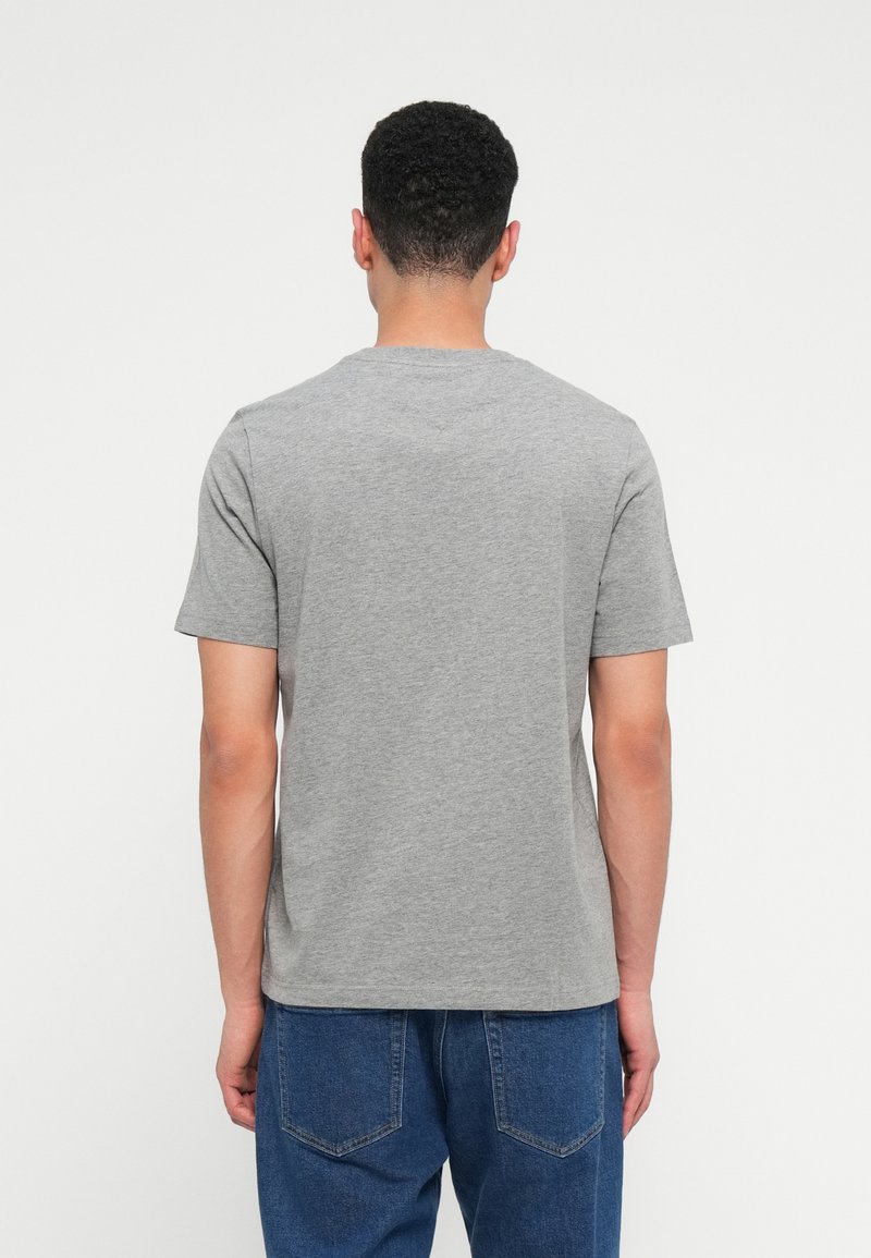 Mand med kort krøllet hår iført en ensfarvet grå T-shirt og blå jeans, vendt væk mod en hvid baggrund.