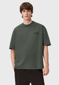 Tummanvihreä puuvillainen T-paita, jossa on pyöreä kaula-aukko ja lyhyet hihat, koristeena hienovarainen musta-logo vasemmassa rinnassa, rento malli.