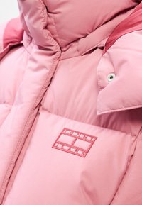 Gros plan d'une doudoune rose clair avec un col montant, un bouton-pression et un logo Tommy Jeans rouge brodé sur la poitrine.