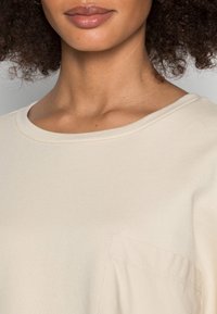 T-shirt beige a coste con scollatura rotonda e una piccola tasca sul lato sinistro. Tessuto morbido e texture.