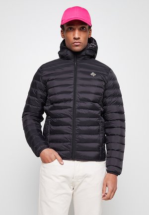 Veste matelassée noire avec motif horizontal, fermeture éclair à l'avant et capuche. Portée avec un pantalon clair, présentant un petit logo sur la poitrine.