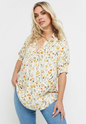 Bluse mit Blumenmuster in Creme mit orange- und gelbtönen, mit V-Ausschnitt, kurzen Puffärmeln und einer lockeren Passform. Kombiniert mit blauen Jeans.