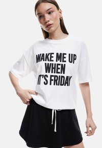 Hvid bomulds t-shirt med sort fed skrift "WAKE ME UP WHEN IT'S FRIDAY." Sammen med en sort snøre nederdel, som præsenterer et afslappet look.