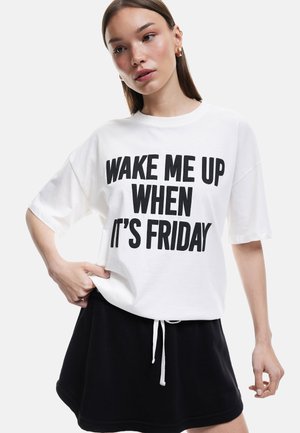 T-shirt en coton blanc avec du texte noir en gras "RÉVEILLE-MOI QUAND NOUS SOMMES VENDREDI." Associé à une jupe noire à cordons, offrant un look décontracté.