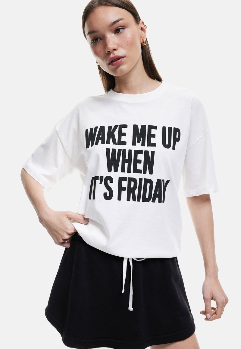 Hvid bomulds t-shirt med sort fed skrift "WAKE ME UP WHEN IT'S FRIDAY." Sammen med en sort snøre nederdel, som præsenterer et afslappet look.