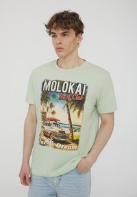 T-shirt di cotone verde chiaro con una grafica vintage da spiaggia raffigurante un'auto, palme e il testo "Molokai Surf Club Pacific Dream."