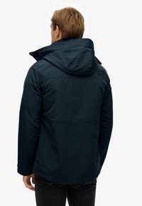 Marineblauwe waterdichte jas met een capuchon, voorzien van een verhoogd logo op de schouder en een gladde, matte textuur.