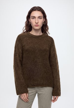 Personne aux longs cheveux bruns portant un pull en laine marron, un pantalon à carreaux et des boucles d'oreilles en or, debout devant un fond clair uni.