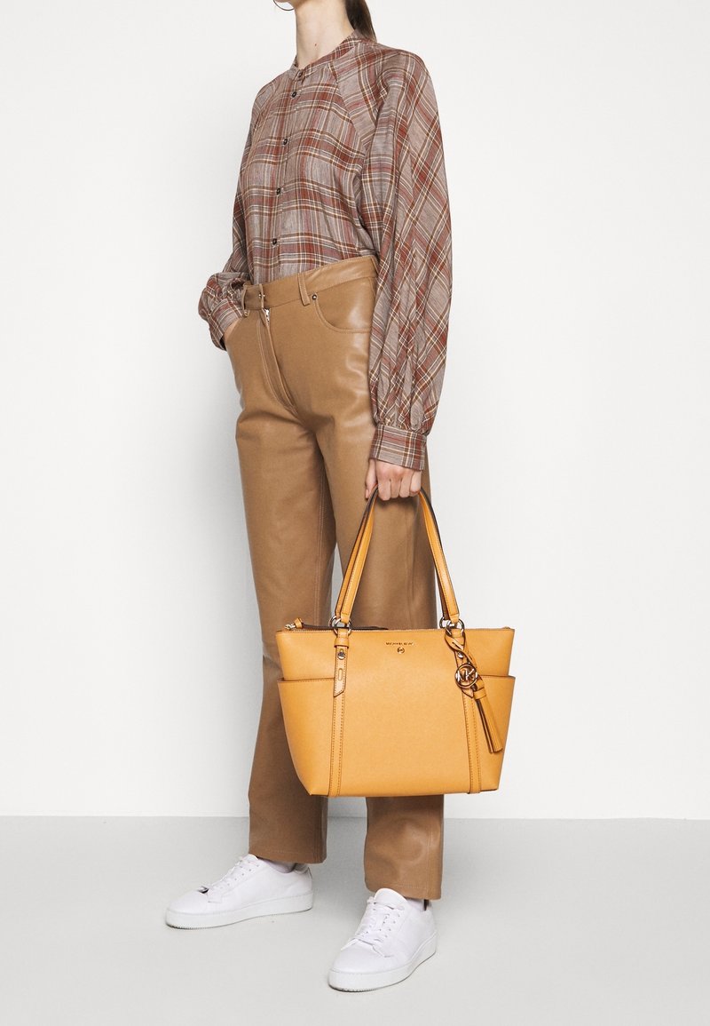 Femme portant un chemisier à carreaux marron et un pantalon en cuir marron clair, tenant un grand sac à main en cuir jaune et portant des baskets blanches.