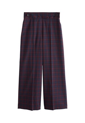 Personne portant un pantalon à carreaux bleu et rouge avec un détail bouton, un pull gris rentré et une main dans la poche.