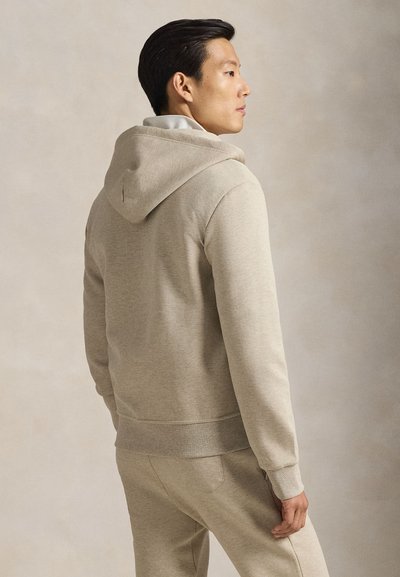 Muž v béžovej mikine s kapucňou a zodpovedajúcich nohaviciach stojí v profile na jednofarebnom béžovom pozadí.