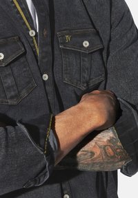 Dunkelblauses Denimhemd mit zwei Brusttaschen, weißen Knöpfen, gelben Stickereiakzenten und hochgekrempelten Ärmeln, die einen tätowierten Unterarm zeigen.
