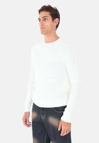 Maglione bianco a maglia con scollo rotondo e polsini a costine, indossato con jeans blu scuro con leggere abrasioni. Texture e vestibilità morbide.