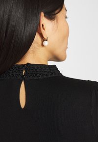 Svart topp med en texturerad spetskrage, en nyckelhålsöppning baktill och ett slätt, åtsittande material. Guldörhängen med pärlzero.