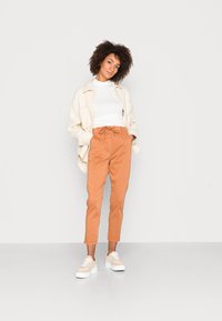 Camisa-jáqueta oversized bege clara com dois bolsos frontais, combinada com calças laranja taper e ténis plataforma bege. Fundo brilhante.