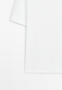 Gros plan d’un t-shirt blanc uni à manches courtes, montrant une manche et une partie du corps sur fond blanc.