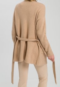 Kvinde iført beige ribbestrikket cardigan med løs bælte, som hun holder i den ene ende, vist fra ryggen mod en ensfarvet baggrund.