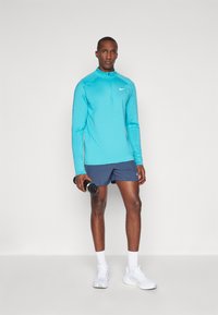 Uomo in piedi con una maglietta sportiva a maniche lunghe turchese, pantaloni corti navy, calze bianche e scarpe da ginnastica, che tiene in mano una bottiglia d'acqua nera con la mano destra.