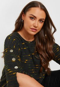 Svart stickad tröja med gula och gröna ränder, med en båtformad halsringning och silverknappar som accent på ärmarna. Mjuk, texturerad tyg.