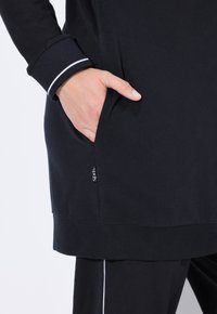 Musta pitkähihaiseen toppi, jossa on sivutasku, ribbed hihansuut valkoisilla yksityiskohdilla ja sileä pinta. Tunnuksessa lukee 'Sports' taskussa.