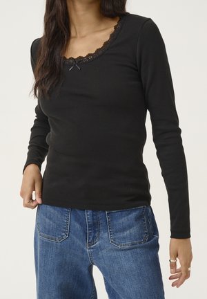 Femme portant un haut noir à manches longues avec bordure en dentelle et petit nœud, associée à un jean en denim bleu, debout devant un fond uni.