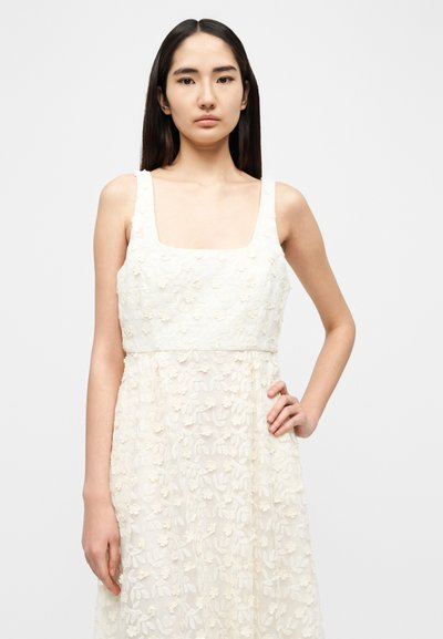 Λευκό φόρεμα με κεντήματα από λουλούδια, τετράγωνο ντεκολτέ, φαρδιές τιράντες και φαρδιά φούστα. Κατασκευασμένο από ελαφρύ, υφασμάτινο ύφασμα με υφή.