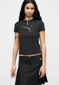 Νεαρή γυναίκα με μακριά σκούρα μαλλιά φοράει μαύρο κοντομάνικο τοπ και μαύρη φούστα με λογότυπα PUMA, στέκεται μπροστά σε απλό φόντο.