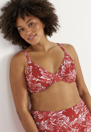 Lächelnde Frau mit lockigem Haar, die ein rotes und weißes florales Bikinioberteil und passende hoch taillierte Bikinihosen trägt und sich an eine weiße Wand lehnt.