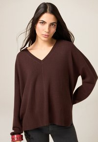 Suéter marrón de cuello en V, ajuste holgado, textura suave, puños y dobladillo acanalados, combinado con jeans oscuros y pulseras apiladas en la muñeca.