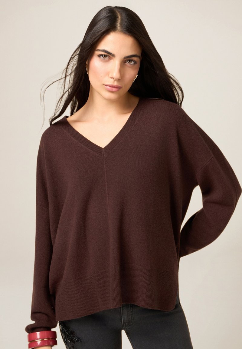 Suéter marrón de cuello en V, ajuste holgado, textura suave, puños y dobladillo acanalados, combinado con jeans oscuros y pulseras apiladas en la muñeca.