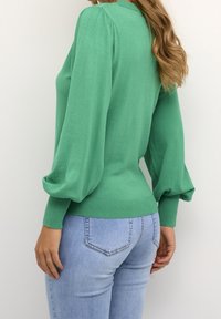 Femme portant un pull vert à manches longues bouffantes et un jean bleu clair ajusté, debout devant un fond blanc uni.