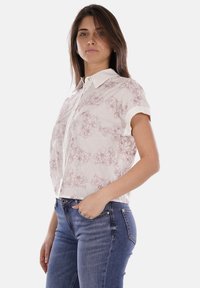 Camicia a maniche corte con bottoni in tessuto leggero, con una base bianca e un motivo floreale in linea rosa. Abbinata a jeans in denim blu.