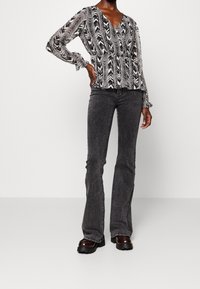 Vrouw in een zwart-witte blouse met lange mouwen en patroon, donkergrijze flared jeans en zwarte platform schoenen, staand met één hand op de heup.
