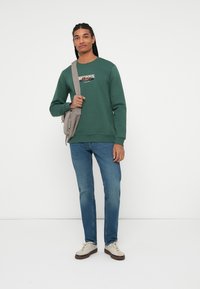 Groene sweatshirt met ronde hals en grafische details op de voorkant, gecombineerd met blauwe jeans en witte sneakers. Grijze schoudertas.