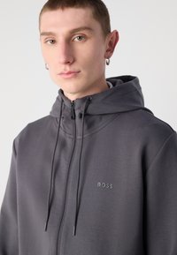 Sudadera con cremallera gris y capucha ajustable con cordón, de textura suave y con un sutil logotipo "BOSS" en el pecho. Cuenta con una cremallera completa.