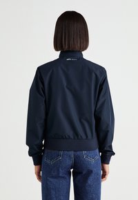 Chaqueta bomber azul marino con cintura y puños acanalados. Presenta un acabado suave y brillante y un logo cerca del cuello. Combinada con jeans de mezclilla.