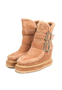 Botas de tobillo de ante marrón con forro de piel de oveja, dos correas anchas con hebillas de cristal y una suela de cuña con un fondo texturizado.