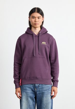 Lilla hoodie lavet af blødt stof, med en frontlomme, justerbare snore og et lille guld-logo på brystet. Bæres sammen med blå jeans.